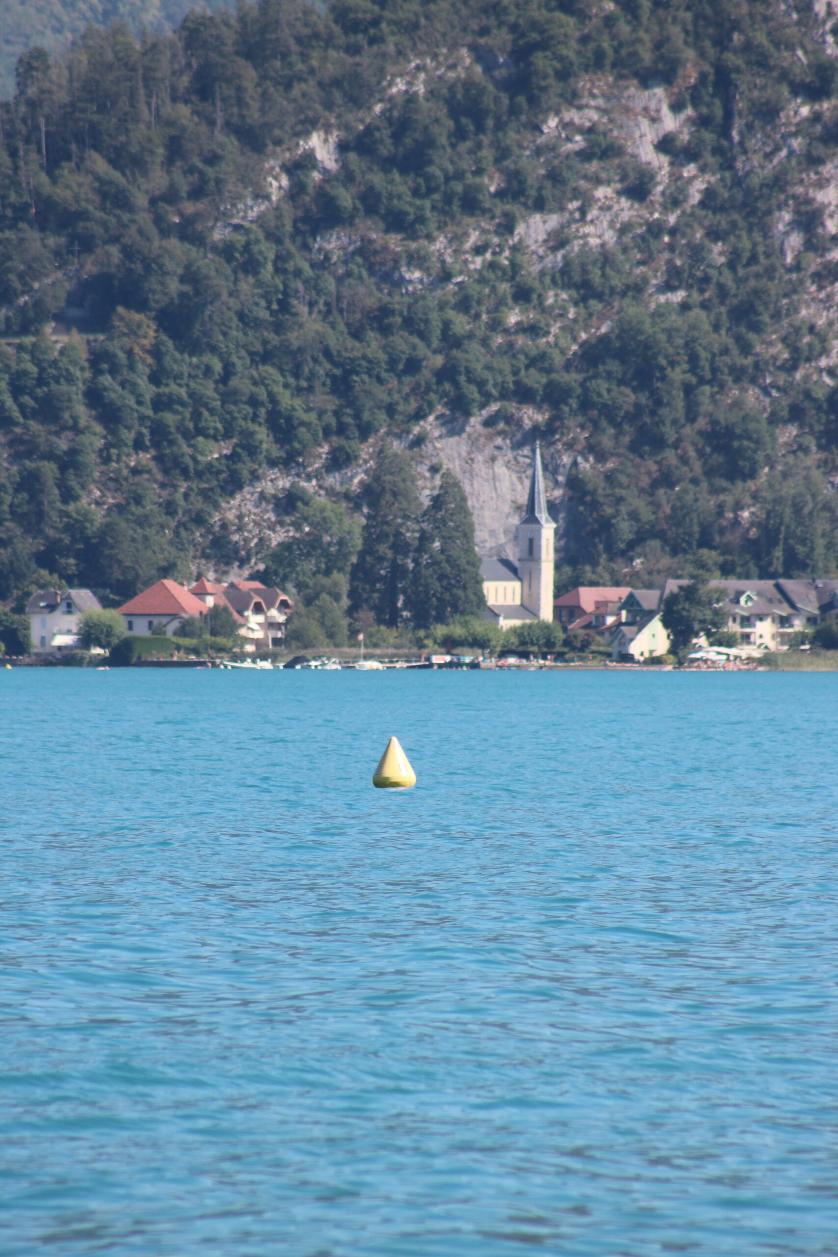 Bouée jaune dans l’eau bleue du lac d’Annecy devant une rive habitée.
