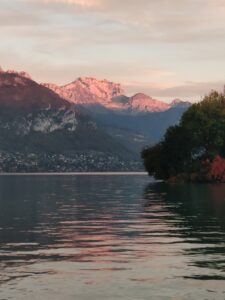 Reflets roses sur le lac d’Annecy devant les montagnes éclairées.