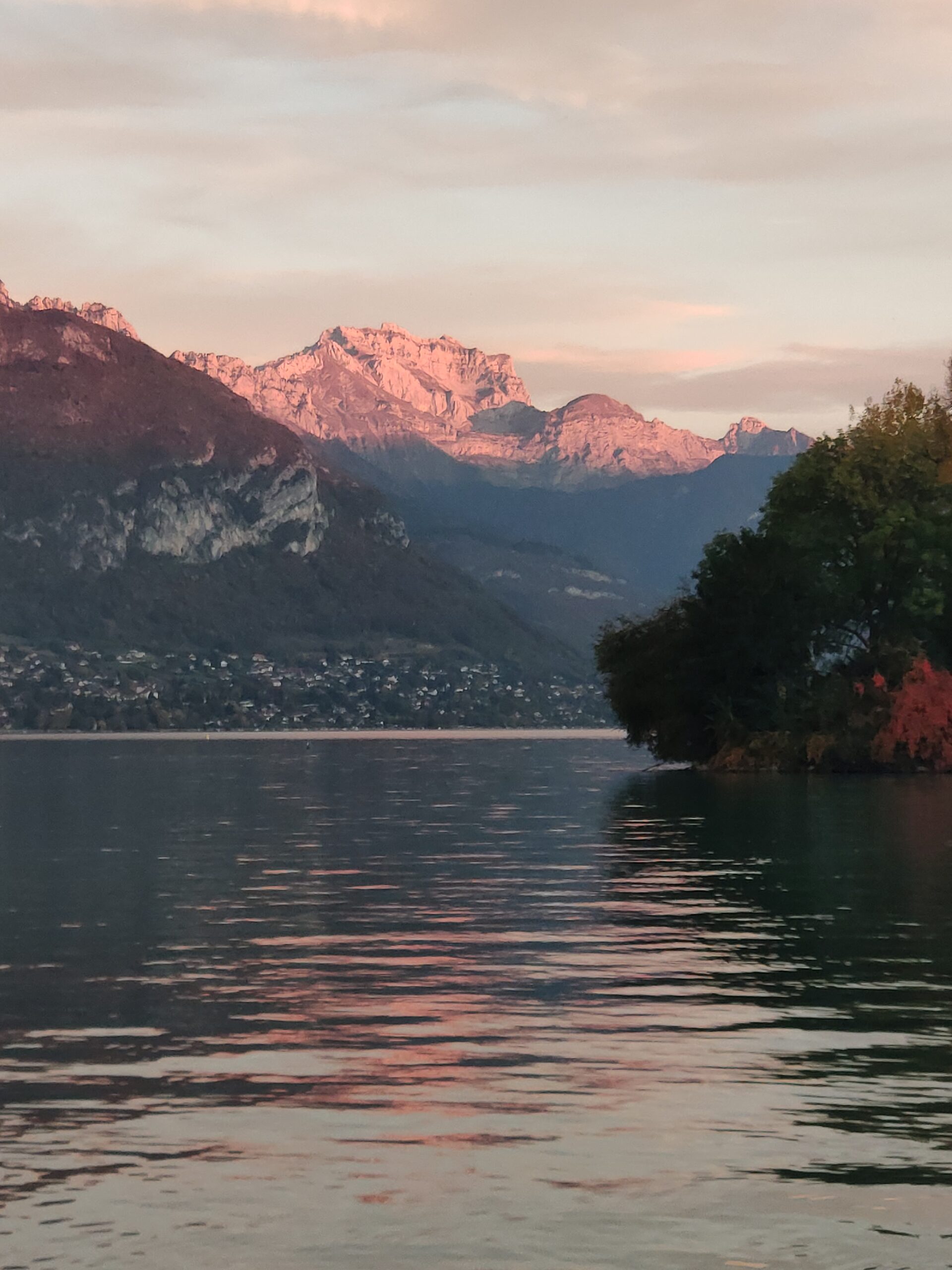 Reflets roses sur le lac d’Annecy devant les montagnes éclairées.